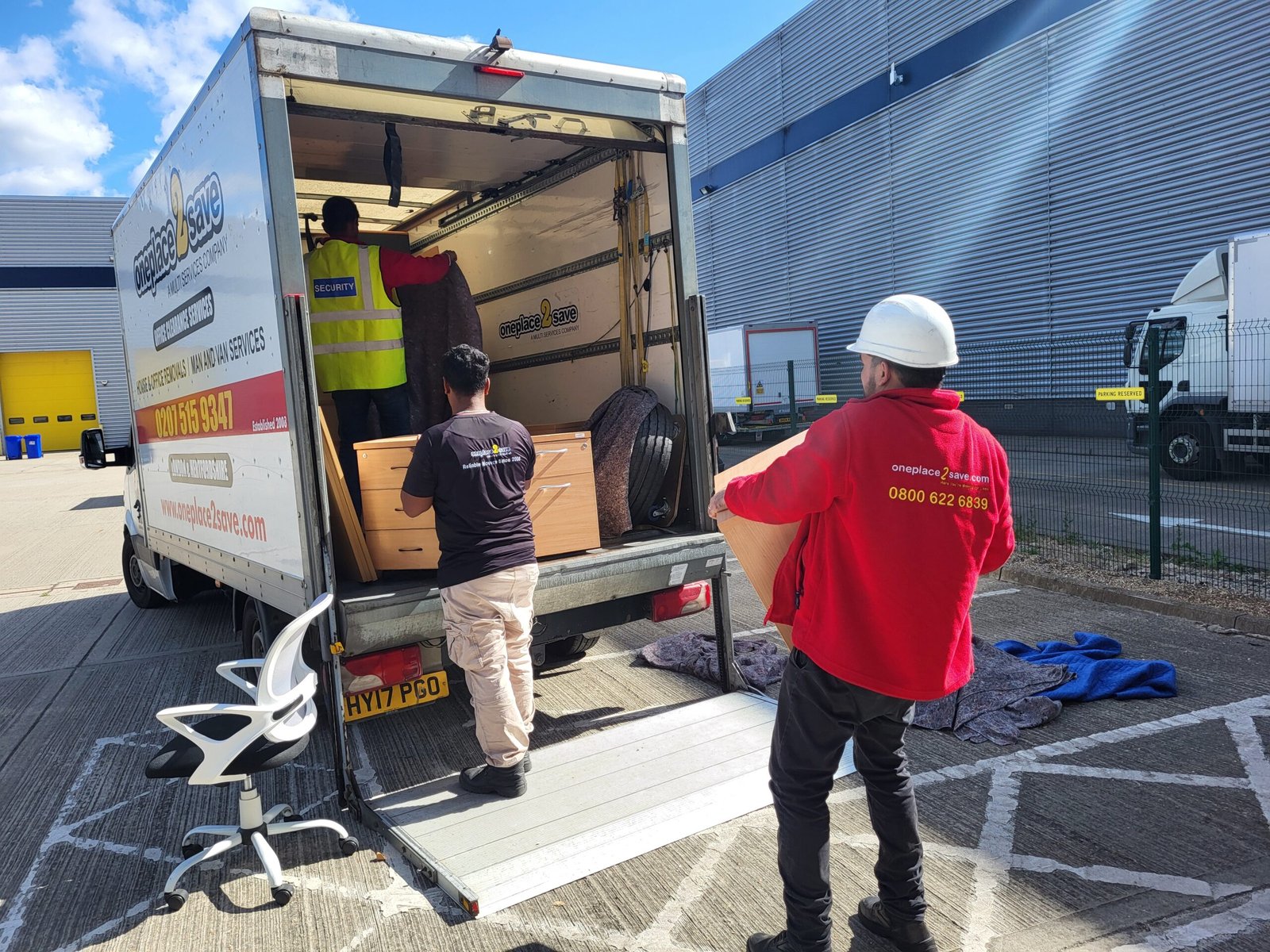 Oneplace2save crew carrying furniture onto a removal lorry at a loading area.