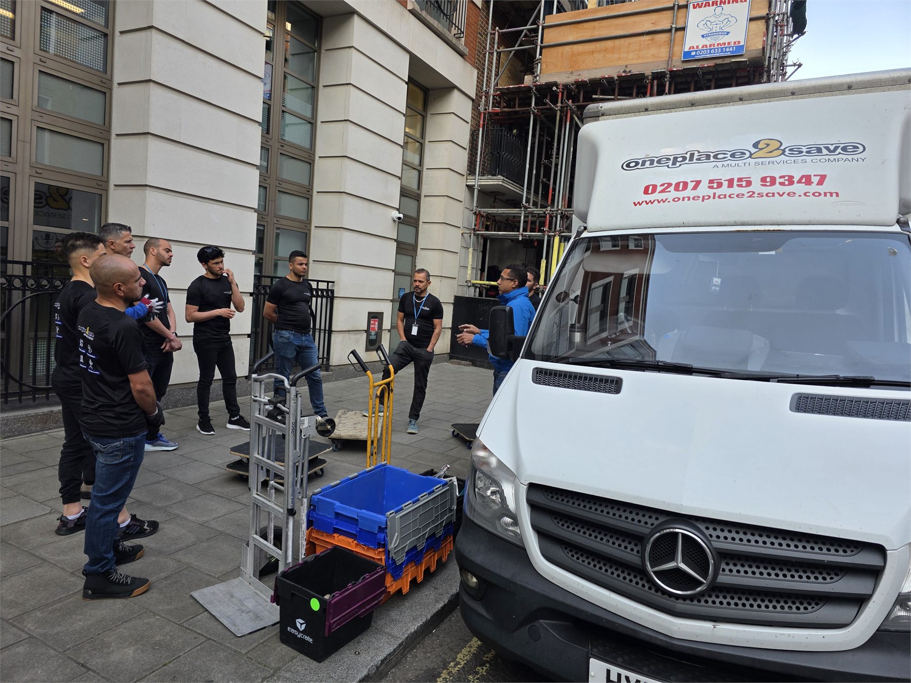 Oneplace2save team beside a branded van, with moving crates and trolleys on the pavement.