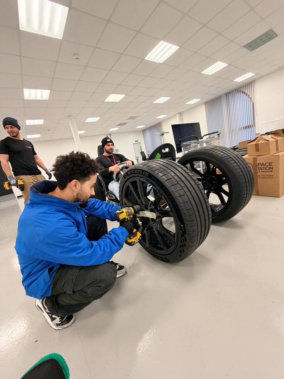 Oneplace2save team member recreating an F1 pit stop with a genuine McLaren wheel and an office-chair rig after the relocation work.
