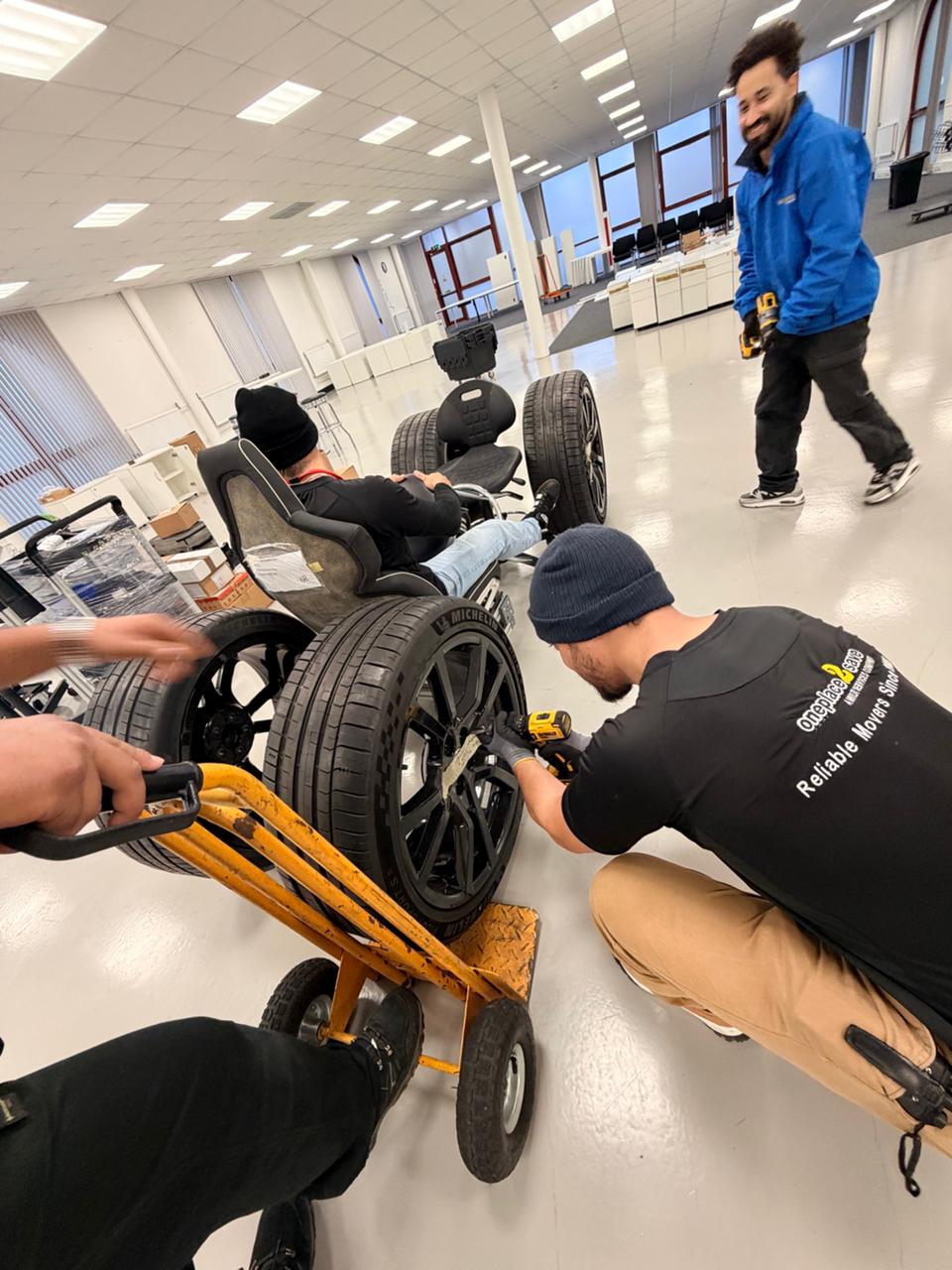 Oneplace2save team recreating an F1 pit stop using genuine McLaren wheels and an office-chair rig after completing relocation work.